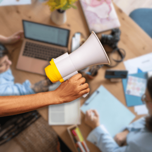 Close-up of a hand holding a megaphone with a creative team working on laptops and documents in the background, symbolizing advertising and promotional strategy.