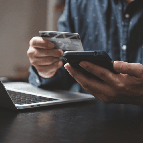 Person holding a credit card and smartphone while shopping online with a laptop in the background, representing e-commerce transactions.